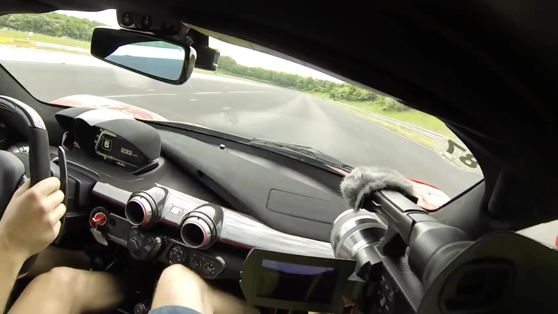 Udsigt fra passagersædet i en Ferrari LaFerrari Aperta på en lige strækning. Displayet viser sjette gear og 233 km/t. Førerens ene hånd hviler på rattet.
